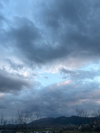 The image displays an outdoor scene in Piperkov Chiflik, Bulgaria, under an overcast sky. The upper two-thirds of the frame are dominated by dense, multi-layered clouds, primarily dark grey to slate blue in coloration, with interspersed patches of lighter blue sky and areas reflecting faint pink or peach light visible through breaks in the cloud cover.

The lower third of the image features a landscape with a dark, undulating mountain range forming the horizon. Below the mountains, a valley contains what appears to be a scattered settlement. In the midground, an open area or field holds multiple parked vehicles, mostly light and dark colored, indistinct due to distance. Bare deciduous tree branches protrude into the frame from the lower left and right, partially obscuring the view. A cluster of blooming trees or bushes with white blossoms is visible in the midground on the right. No individuals, specific actions, or interactions are discernible in the image.