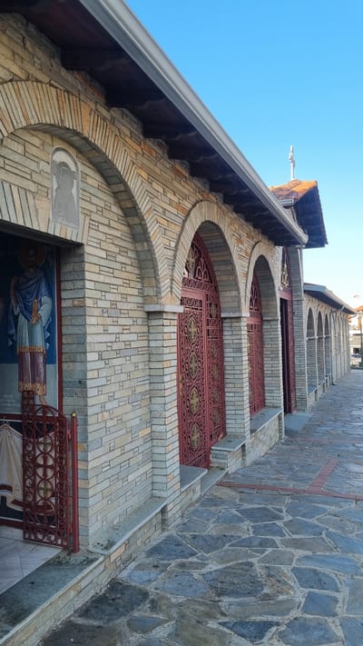 The image shows the exterior of a church in Litochoro, Greece, on a clear day. The church is constructed with light-colored stone bricks and features a series of arched doorways with ornate red metal gates. A fresco of a religious figure is visible on the left side of the building, next to an entrance. The ground in front of the church is paved with irregular flagstones. The architecture suggests a traditional Eastern Orthodox style, common in the region. The sky is a vibrant blue, and the sun casts subtle shadows on the stone facade, indicating it is likely morning or late afternoon. No people are visible in the immediate vicinity. The overall impression is one of a well-maintained religious building in a Mediterranean setting.