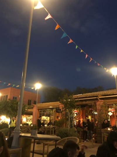 A string of colorful triangular flags stretches across a dark blue night sky, illuminated by two streetlights. The flags are strung between a white pole on the left and a building on the right. The building is a two-story structure with a pink facade and a series of windows. The building is partially obscured by a leafy tree. The scene is located in Palaio Faliro, Greece, at the coordinates 37.929 latitude and 23.685 longitude. The building is a restaurant and has a sign that says "Cru" on the upper level. People are sitting at tables and enjoying the evening atmosphere. There are multiple lights in the area that illuminate the scene.  The light from the streetlights and building lights illuminate the space, providing a warm and inviting ambiance.  A white table with a white top and a white base stands on the ground, just under the streetlight on the right.