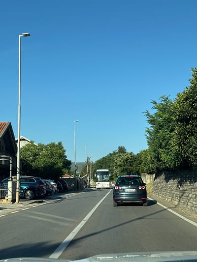 The image shows a sunny day in the city of Bijela, Montenegro, likely along a road leading out of town. A dark grey Volkswagen Golf is the prominent subject in the foreground, driving on the right side of the road. The car's license plate reads "TV BF 074". Ahead of it, a white bus with "CARINE" written on its side is also driving in the same direction. Several other vehicles are parked on the left side of the road, including a dark blue SUV. The road is asphalt with white lane markings.
To the left of the road, there are buildings with tiled roofs and lush green trees. A tall street lamp stands prominently on the left side of the road. To the right of the road, a stone wall covered in greenery lines the street, with more trees further back, suggesting a hilly or mountainous terrain. The sky is a clear, vibrant blue with no visible clouds. The scene appears to be captured from the perspective of a vehicle also traveling on the road.