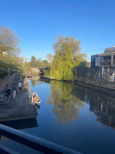 The image shows a canal scene in Kolding, Denmark on a sunny day. The canal reflects buildings and a large weeping willow tree. A group of people is gathered on a wooden dock or platform at the edge of the canal. Some people are seated on the steps of the dock. A small boat is pulled up on the dock near the group. A bridge or sluice gate structure spans the canal further in the distance. The sky is clear and blue.
