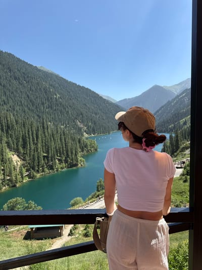 The image, oriented vertically, shows a woman from behind, leaning over a railing and looking out at a scenic mountain lake. She is wearing a white short-sleeved shirt, white linen pants, and a beige baseball cap. A pink scrunchie is visible in her hair, and she has a light-colored bag slung over her shoulder.
The background reveals a stunning natural landscape. A vast, turquoise lake stretches out below, flanked by steep, densely forested mountains. The trees appear to be coniferous, creating a textured, dark green carpet on the slopes. The sky is a clear, bright blue, suggesting a sunny day. The time of day appears to be midday, given the bright sunlight casting shadows.
The woman's posture suggests she is admiring the view, perhaps contemplating the grandeur of the natural surroundings. The setting is clearly outdoors, likely at a viewpoint or balcony overlooking the mountainous terrain and lake. In the distance, a small wooden structure with a green roof is visible near the lakeshore. The overall mood is one of peace and appreciation for nature's beauty. No visible text is present in the image. The location is identified as Kurmeti, Kazakhstan.