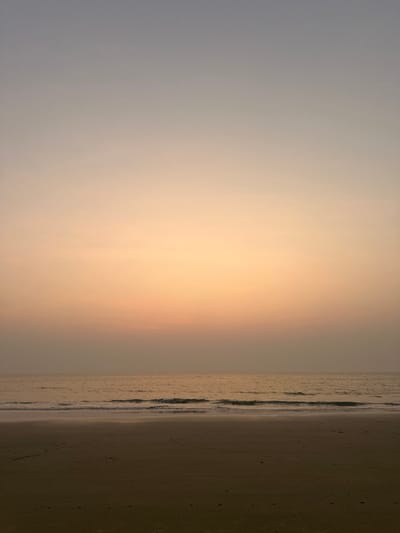 The image displays a coastal scene at Betalbatim, India, characterized by a vast, empty expanse. The upper two-thirds of the frame consists of the sky, exhibiting a subtle gradient from light grey-blue at the zenith, transitioning through pale orange-yellow, to a soft, hazy pinkish-orange just above the horizon line. No distinct clouds are visible. The middle section, approximately one-quarter of the image, depicts the sea, reflecting the warm hues of the sky with a muted golden-brown coloration. Small, low-amplitude waves are breaking close to the shore, indicated by subtle lines of white foam. The bottom quarter of the image is comprised of a flat, dark brown sandy beach, appearing wet along the waterline. No individuals, animals, structures, or objects are present within the frame, and no actions or interactions are depicted.