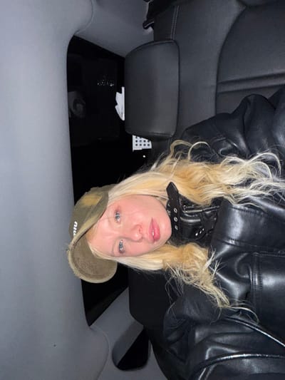 A woman with blonde hair and blue eyes wearing a black leather jacket and a Miu Miu baseball cap looks directly at the camera. She is positioned in the backseat of a car with a dark interior. The car's headrest is visible on the left, and the floor is obscured by shadows. A partial view of the ceiling can be seen.