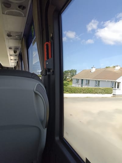 The image is taken from the inside of a bus in Kincasslagh, Ireland, looking out the window. In the foreground, a grey bus seat is visible. The window shows a bright blue sky with scattered white clouds. Outside the window, a light-colored residential house with a brown tiled roof is visible. The house has multiple windows and a chimney. A neat green hedge runs along the front of the property, with a white wall below it. The ground in front of the house appears to be paved or a light-colored road. There is also a red emergency handle mounted on the interior of the bus frame next to the window.