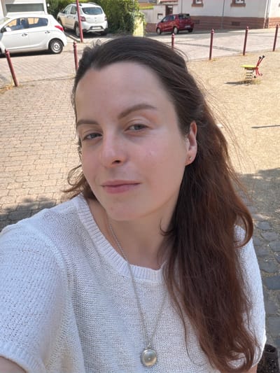 A woman with long, brown hair and fair skin is captured in a close-up, selfie-style shot. She is wearing a white, knitted sweater and a silver necklace with a circular pendant. Her expression is neutral, and her eyes are looking towards the camera.
In the background, a paved area with a cobblestone pattern is visible, suggesting an outdoor setting. Sunlight casts shadows on the ground, indicating it is daytime. A red car is parked in the distance, and a building with brown accents is partially visible. Further in the background, there are hints of greenery and what appears to be a playground structure with red and yellow elements.
The image seems to be taken on a sunny day. The scene suggests a casual outdoor environment, possibly a street or a public area. No visible text is present in the image. The location context provided is Monsheim, Germany.