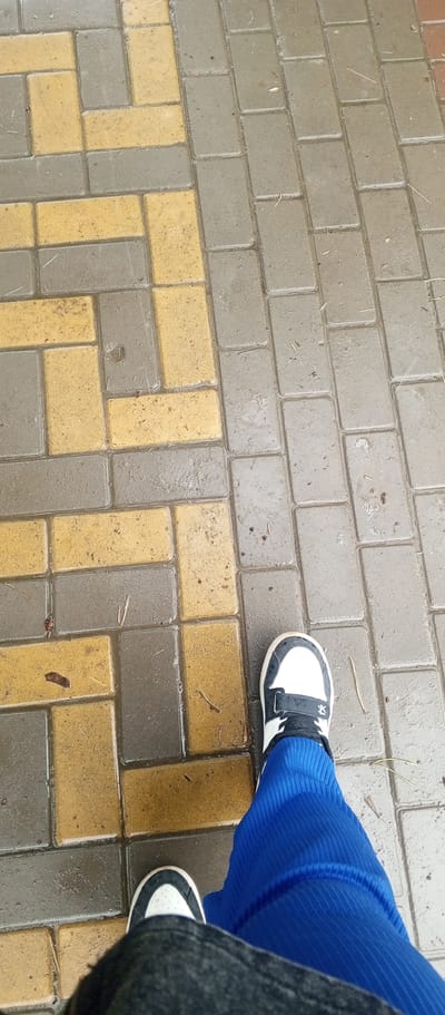 The image shows a perspective view of a person's legs and feet standing on a wet, patterned pavement. The pavement is composed of interlocking rectangular blocks, alternating in color between gray and yellow. The person is wearing dark pants and a blue, ribbed skirt or wide-legged trousers. On their feet are white sneakers with black accents and laces. The overall environment appears to be outdoors, possibly a street or walkway in the city of Zhirnovsk, Russia, given the presence of wet pavement, suggesting recent rain. There are small pieces of debris, like twigs and leaves, scattered on the pavement.