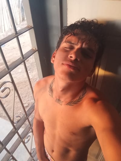 A shirtless young adult male with dark, curly hair is leaning against a wall, taking a selfie. He has a tattoo of a laurel wreath around his neck, with the number "2005" visible below it on his chest. He is wearing patterned underwear. Behind him, to the left, is a barred window with a sandy ground visible outside, and some wooden structures. To the right, the wall is illuminated by warm light, suggesting the sun is setting or rising. The overall impression is of an informal, personal moment captured in a somewhat rustic environment.
