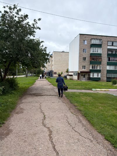 A person in a dark blue jacket walks away from the camera on a cracked concrete sidewalk. The sidewalk is bordered by a strip of green grass, and beyond that are multi-story buildings. The buildings appear to be residential apartment blocks, with several windows visible. One building is lighter colored and features a white, corrugated siding. Another is constructed of brick and has green trim around some of its windows. Trees and greenery are also present in the scene. The sky is overcast and cloudy. The time of day appears to be daytime. There is some visible text on a sign attached to one of the buildings, reading "ПЕРМСКИЙ КАРЕ" and another sign below it which is partially obscured.