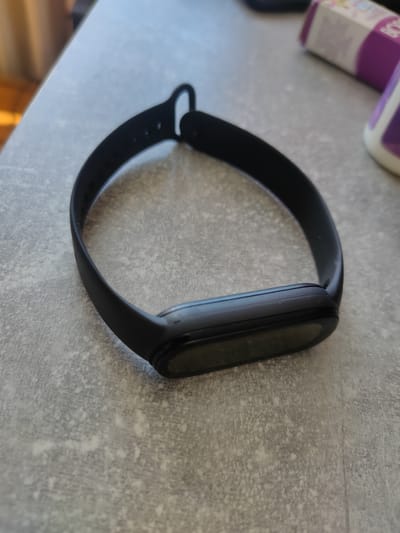 A black fitness tracker with a dark display lies on a speckled grey countertop. The tracker is positioned in the center of the frame, with its strap curved around it, suggesting it is not currently being worn. The background is out of focus, but hints of a light-colored surface and what appear to be product packaging are visible. No people or activity are present in the image.