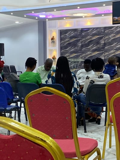 This image depicts an indoor gathering in Gbawe, Ghana. Seven individuals are visible, primarily seated. In the immediate foreground, two red upholstered chairs with gold metallic frames are out of focus.

From left to right, in the midground: A person in a red head covering is partially visible, seated. A woman with dark, short hair, wearing a green top, is seated with her back partially to the camera, looking towards the man next to her. A bald man in a light green collared shirt is seated, leaning forward and looking down. A woman with long dark braids, wearing a patterned kaftan-style top (blue, white, yellow), is seated with her back partially to the camera. A man in a white long-sleeved top with a dark graphic design is seated, looking down. To his right, a woman in a white and black patterned top is seated, holding an infant dressed in a blue garment. The infant's face is not visible.

The seating consists of dark chairs occupied by the subjects, with additional rows of dark chairs visible behind them. The room features a decorative background wall. The central portion of this wall has a dark grey and white marble-patterned surface, framed by white architectural paneling above. This paneling includes recessed yellow lighting and two alcoves containing vases with red artificial flowers and green foliage. The ceiling is white, with multiple recessed lights displaying white and purple-blue illumination, along with a central circular light fixture. A large black speaker is partially visible on the far left. A section of a screen or display is visible high on the far right wall. The floor is a light-colored, reflective surface.