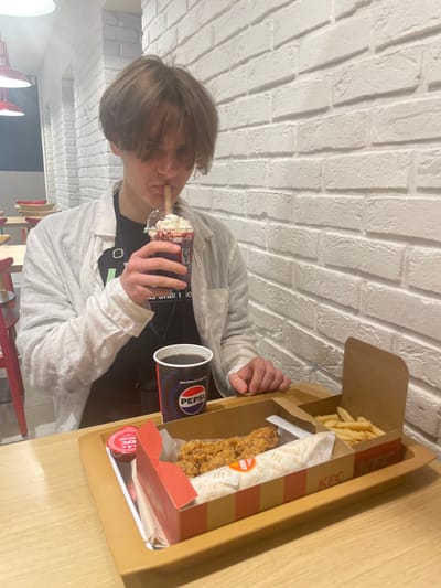 The image depicts a young man sitting in a fast food restaurant in Jiříkovice, Czechia (49.184, 16.761). He is wearing a white linen shirt over a black t-shirt and is drinking a milkshake through a straw. In front of him is a tray containing a KFC meal consisting of fried chicken, fries, and a small container of sauce. Next to the milkshake is a cup of Pepsi. The man's right hand is holding the milkshake and his left hand is resting on the tray. The restaurant is brightly lit and has white brick walls. The table surface is made of wood. The table is empty except for the tray and the man's belongings. The man is looking down at his milkshake while he drinks. The image is focused on the man and his food. It captures a moment of everyday life in a fast food setting.