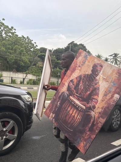 A man stands beside a dark car, holding a large, framed painting of a man playing a drum. The scene is located in Falomo, Nigeria, as indicated by the urban environment featuring lush green vegetation and palm trees. The man stands on the side of a paved road with traffic markings. Overhead power lines are visible against a cloudy sky. The car is positioned on the road.