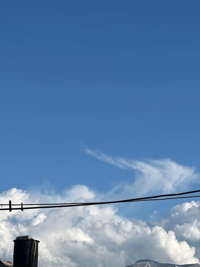 The media file is a photograph taken from a low angle, looking upwards towards a bright blue sky. The sky is mostly clear, with a few wispy white clouds scattered across it. On the right side of the frame, there are large, puffy white clouds, suggesting fair weather. Two thin, dark wires run vertically through the center of the image, intersecting with a ladder-like structure. To the far right, a dark, cylindrical object, possibly a part of a building or industrial equipment, is partially visible. The lighting indicates it is daytime, with the sun high in the sky. There is no discernible activity or event taking place, and no text is visible. The overall impression is one of a clear, open sky with some man-made elements intruding into the natural scene. The location is specified as Tobyl, Kazakhstan.