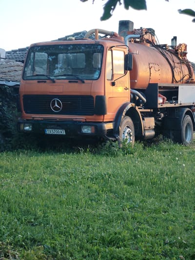 The image depicts an orange Mercedes-Benz truck with a tank, parked on a grassy area in Kochmar, Bulgaria. The truck is the primary subject, with its front grill prominently featuring the Mercedes-Benz logo. A license plate reading "TX 5700 AT" is visible on the front of the truck. The tank appears to be a septic or vacuum truck, indicated by the piping and hoses attached to it. The background shows a building with a tiled roof, possibly a house or structure in Kochmar. The environment is a field of green grass, with some leaves from an overhanging tree visible at the top of the image. The lighting suggests it is either early morning or late afternoon, with a warm glow illuminating the truck. There are no people visible in the image, and no actions are taking place. The scene is static and captures the truck in its parked state.
