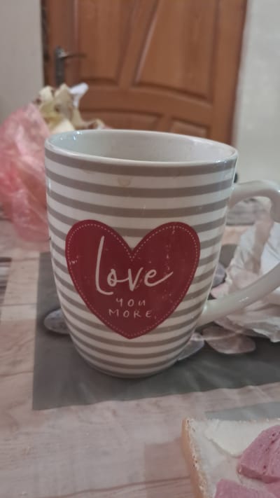 A close-up shot shows a mug with a heart design on a table. The mug is gray and white striped, with a red heart in the center that says "love you more". A piece of toast with white spread and pink pieces is in the lower right corner of the frame. The background shows a wooden door and a blurred pink plastic bag. The scene is likely a home interior in Krynychky, Ukraine.