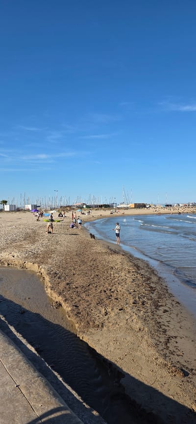 The image shows a beach in Canet d'en Berenguer, Spain. To the left is the open blue sky, transitioning to the beach, filled with beachgoers. There are numerous small boats lined up along the shore, suggesting the presence of a marina nearby. A person stands near the water's edge. A shadow is present at the bottom right corner. The beach is littered with seaweed.