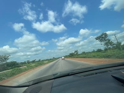 The image depicts a view from the driver's seat of a vehicle traveling on a paved road through a rural landscape in Aciol, Nigeria. The road stretches into the distance, with two lanes visible and a car ahead. The surrounding environment is characterized by lush green vegetation and scattered trees, with a clear blue sky dotted with white, fluffy clouds overhead. The time of day appears to be late morning or early afternoon, given the bright sunlight and the angle of the shadows. The weather is fair and sunny, suitable for travel. The scene conveys a sense of movement and journey through a serene, natural setting. There are no visible people or text in the image.
