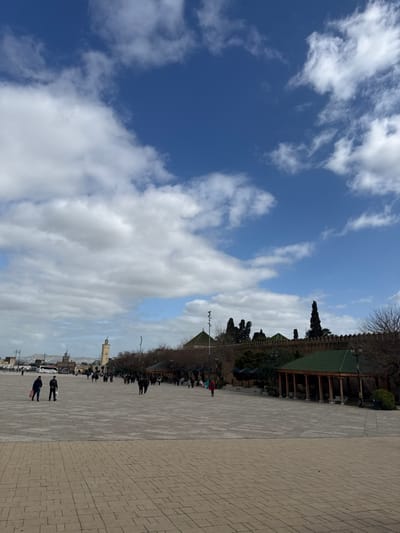 This image depicts the expansive Mechouar plaza in Fes Jdid, Morocco, under a partially cloudy blue sky. The foreground and midground are dominated by a large, light-colored paved area, possibly stone or concrete, laid in a grid pattern.
In the midground, to the right, a long, tan defensive wall with crenellations extends horizontally. Behind this wall, traditional Moroccan architecture featuring multiple green-tiled domed roofs and dark green cypress trees is visible. Along the plaza side of the wall, several open-sided pavilions with green-tiled roofs and light-colored columns are present. Bare deciduous trees are planted along the wall.
In the distant left background, a tall, slender, light-colored minaret with a square base stands prominently. Further beyond, other light-colored buildings with flat roofs and a distinct arched gateway are discernible, along with vehicles, including at least one bus, on a road. Distant hills are faintly visible on the horizon.
Approximately 20-30 individuals are scattered across the plaza, predominantly walking. In the foreground, left of center, two individuals walk towards the viewer; one wears a dark jacket and light pants while carrying a light-colored bag, and the other wears dark clothing. No specific interactions or detailed activities are clearly identifiable among the distant figures. The scene is illuminated by natural daylight.