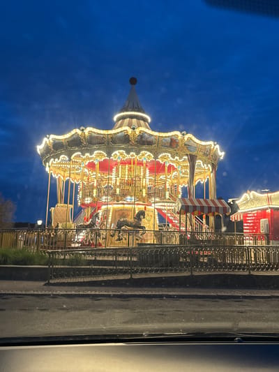 An illuminated carousel is situated outdoors at dusk in Gdańsk, Poland. The carousel is elaborately decorated with gold trim and red accents, and it is lit by numerous warm white lights. The carousel is circular and has multiple levels, with horses and ornate seating visible. A red and white striped awning is visible near the entrance to the carousel. To the right of the carousel, a smaller red structure with a similar awning is also illuminated. The carousel is enclosed by a decorative metal fence. Beyond the fence, there is grass and a lamppost. The ground in front of the carousel appears to be wet, possibly from recent rain. The sky above is a deep blue, indicating twilight. The image is taken from the perspective of someone looking out from a vehicle, as suggested by the dark dashboard-like element at the bottom of the frame.