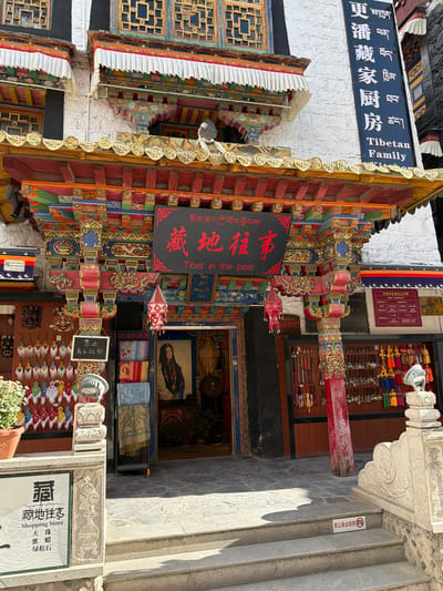 This image presents a vibrant street scene featuring the entrance to a traditional Tibetan-style building, likely in Chengguan District, China. The structure is characterized by richly carved and painted wooden elements in a palette of reds, blues, greens, and gold, adorned with intricate patterns.
Above a shop entrance, a prominent black sign reads "藏地往事" (Zàngdì wǎngshì), translated as "Tibet in the past," accompanied by Tibetan script "སྔོན་ཆད་ཀྱི་བོད་ལྗོངས།". To the left, a large blue sign on a white wall displays "藏家厨房" (Zàngjiā Chúfáng), translated as "Tibetan Family." The entrance itself leads to an area displaying various merchandise, including colorful textiles, possibly shawls or scarves, and smaller decorative items. Inside, a framed portrait is visible.
A stone pillar on the right side of the frame features a sign for a "藏地往事 Shopping Store," listing items such as "天珠" (Dzi beads), "蜜蜡" (Amber), "绿松石" (Turquoise), and "尼泊尔披肩" (Nepalese Shawl). Two distinct white signs with a red circle and slash explicitly state "禁止商业拍照" (No commercial photography), indicating restrictions on photography for commercial use. A smaller red sign high on a wall also mentions a "文物安全责任公示牌" (Cultural Relics Safety Responsibility Public Notice Board).
The scene is illuminated by bright daylight, casting noticeable shadows on the stone steps and ground, suggesting a clear, sunny day. The combination of traditional architecture, diverse merchandise, and multilingual signage points to a cultural and commercial hub.