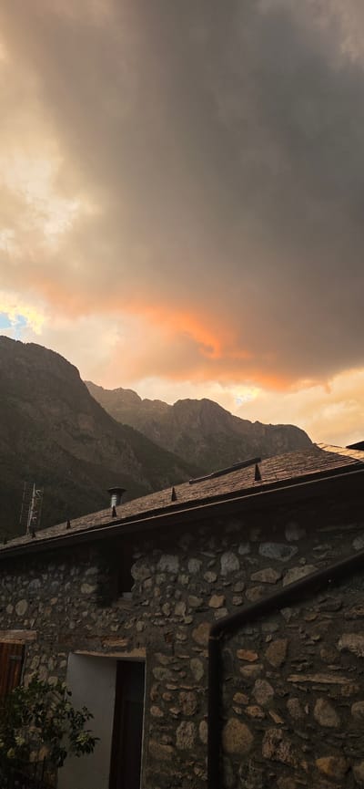 The image captures a dramatic sky over a mountain landscape and a stone building. The sky is filled with heavy, dark clouds, with a vibrant streak of orange and yellow light breaking through towards the horizon, suggesting a sunset or sunrise. In the mid-ground, a range of imposing mountains is visible, their peaks partially obscured by the atmospheric conditions. In the foreground, a rustic stone building with a dark, tiled roof occupies the lower portion of the frame. The roof has several prominent spikes along its edge, and a metal chimney cap is visible. A television antenna is mounted on the roof to the left. The building's wall is constructed of rough-hewn stones of various sizes. To the left of the building, some greenery and the edge of a wooden shuttered window are visible. The overall impression is one of natural beauty and rustic charm in a mountainous setting. The location is identified as Benasque, Spain.