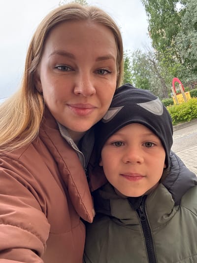 A woman and a young child are in a close selfie outdoors. The woman has blonde hair and is wearing a brown jacket. The child is wearing a black hooded hat with white designs and a green jacket. They are both looking at the camera with a slight smile. The background includes trees, a fence, and a playground structure, suggesting a park or recreational area. The location is in the city of Fedorino, Russia.