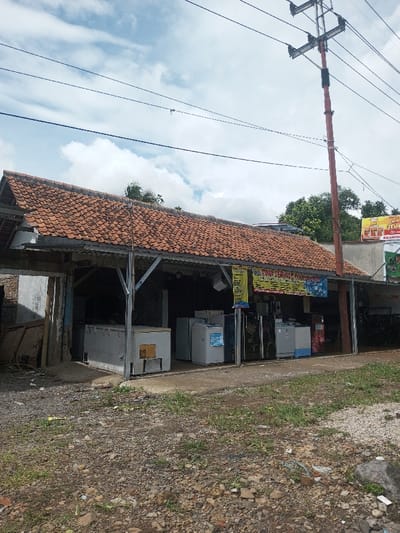 A roadside shop is under a terracotta-tiled roof, supported by a metal frame. The shop displays an assortment of used refrigerators and freezers. A utility pole with overhead wires stands nearby. The ground in front of the shop is gravelly with sparse vegetation. Banners hang above the refrigerators, and a commercial sign is visible on the adjacent building. The sky above is partly cloudy.