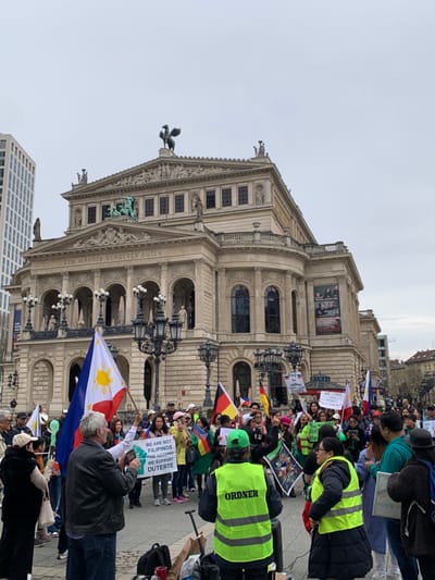 The scene takes place in front of the Alte Oper (Old Opera House) in Frankfurt am Main, Germany (coordinates: 50.115, 8.672). A large group of people are gathered in front of the building, holding signs and flags in a demonstration. The flags include the Philippine flag and the German flag. Many of the signs are written in English, displaying messages such as "We are not Filipinos for Duterte, we support the Philippines", and "You stand with the oppressor". The protesters are a mix of men and women of diverse ages and ethnicities. Some are wearing vests that say "Ordner" (German for "organizer") on them. There are police officers present, wearing reflective vests and standing near the protesters. The environment is predominantly paved with concrete, with the building in the background. The weather is overcast and cloudy. The building is a large, ornate structure with columns, statues, and a dome. There are trees and other buildings visible in the distance. Many of the protesters are facing the camera, while some are looking toward the building. There are various other objects scattered around the scene, including backpacks, umbrellas, and cameras. The overall atmosphere is one of tension and activism.