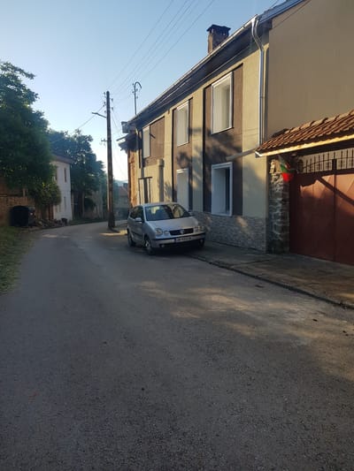 The image depicts a street scene in the city of Lomets, Bulgaria. In the foreground, a silver Volkswagen Polo is parked on the right side of a paved road. The car has a license plate that reads "OB 9055 BC". To the right of the car is a light-colored building with a dark brown metal gate and a tiled roof. A Bulgarian flag is attached to the wall above the gate. The building has stone accents around the gate area and a chimney on the roof. The road stretches into the distance, lined with trees and houses. On the left side of the road, there is a black trash bin and some wooden logs. Overhead, power lines crisscross the sky, supported by a wooden utility pole. The scene is illuminated by sunlight, casting shadows on the road and buildings, suggesting it is either early morning or late afternoon. The sky is clear and blue.