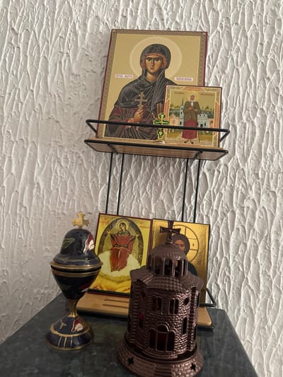 The image displays a collection of religious items arranged on a dark, possibly granite or marble, surface and a two-tiered wire-and-wood shelf unit against a textured white wall, located in Zankovići, Montenegro.
On the upper shelf, a large Orthodox icon of Saint Paraskeva, depicted in dark robes holding a cross, stands upright. Its Cyrillic inscription reads "ПРВ. МАТИ ПАРАСКЕВА." In front of it, a smaller icon portrays an elderly female saint with a staff, situated before a cityscape. A small green and gold Orthodox cross on a silver base is positioned directly in front of the smaller icon.
On the lower surface, to the left, is a dark blue and gold enameled censer with a domed, perforated lid topped by a gold Orthodox cross. It features red and green floral designs. To the right, a dark brown, miniature architectural structure resembling an Orthodox church, complete with a dome and cross, serves as a decorative item or holder. Behind these objects, two more icons are partially visible: one on the left depicts a female saint in red robes with outstretched arms, seated on a cloud against a golden background; the other, largely obscured by the church-like structure, shows a portion of a male saint with a halo and beard against a golden background.