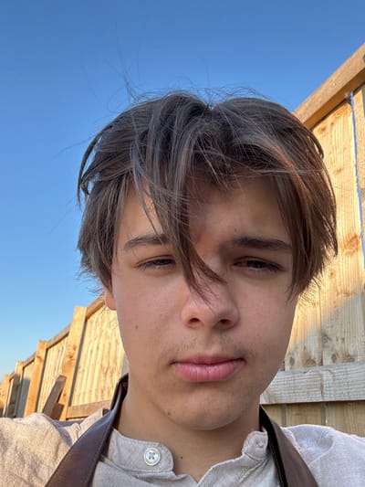 A close-up portrait, taken outdoors in Northallerton, United Kingdom, features a young male subject. The individual has disheveled, medium-length light brown hair that falls across his forehead, partially obscuring his right eye. His eyes are dark, and faint facial hair is present above his upper lip and on his chin. He wears a light-colored, possibly linen, button-up shirt with a visible light-toned button on the placket. A dark brown strap, consistent with an apron or satchel, is visible over his left shoulder. The background on the left and upper portion consists of a clear, bright blue sky. On the right and extending into the lower left, a light-toned wooden fence or structure, composed of vertical timber planks, is visible, illuminated by a warm, direct light source. The subject maintains a direct, neutral gaze towards the camera.