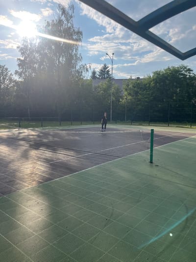 In Kobryn, Belarus, a person is engaged in an activity on a tennis court. The scene is set outdoors on a sunny day, with a bright sun casting lens flares and highlighting the green, textured surface of the court. The court is marked with lines, and a tennis net stretches across its width, supported by a green pole. Trees and foliage surround the court, with a glimpse of buildings in the background. The sky is a clear blue with scattered white clouds. The lighting suggests it is late afternoon or early evening due to the long shadows cast on the court. The overall atmosphere appears peaceful and recreational.