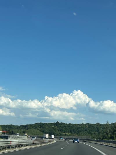 The image depicts a highway scene with cars and trucks traveling under a bright blue sky with scattered white clouds. The road curves to the right, bordered by metal guardrails. Beyond the highway, a dense, green forest covers rolling hills. The context of Baliovtsi, Bulgaria, suggests this is a view of a major roadway likely connecting or passing through the region. The image is captured from a moving vehicle, as indicated by the perspective and the motion blur present in some elements. There are no visible people in the image. The overall scene is characteristic of a sunny day on a highway in a rural or semi-rural area.