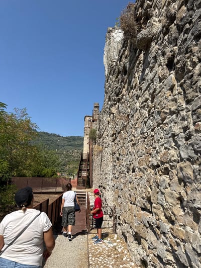 In Dolceacqua, Italy, three people are walking on a gravel path alongside a rough stone wall. The person in the foreground, viewed from behind, wears a white t-shirt and a black baseball cap. Ahead of them, a person in a white t-shirt and camouflage shorts walks away, carrying a dark blue bag. To the right, a person in a red t-shirt and black shorts, wearing a red baseball cap, stands beside a metal gate. Behind the people, a rusty metal structure with stairs ascends alongside the stone wall, and further back, a green hillside with scattered buildings and trees is visible under a clear blue sky.
