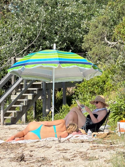Two individuals are relaxing on a sandy beach in Hampton Bays, United States of America, under a striped beach umbrella. The person in the foreground is lying face down on a towel, wearing a turquoise bikini bottom. Their back is to the camera, and their legs are crossed. The second person is sitting in a lounge chair, facing left, and is reading a magazine. This individual is wearing a straw hat, sunglasses, and a dark-colored bathing suit top with a patterned sarong. A water bottle and a straw bag are on the sand near the chair. Wooden stairs are visible to the left, leading upwards behind the umbrella. The background consists of dense green foliage and trees, suggesting a natural coastal environment. The lighting indicates a sunny day.