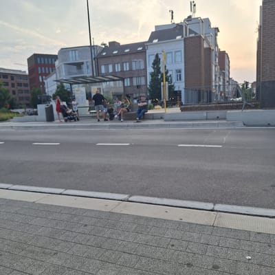 The image shows a street scene in Mechelen, Belgium. In the foreground, a grey tiled sidewalk is visible, bordered by a grey curb and an asphalt road with white lane markings. In the middle ground, several people are gathered on a sidewalk or bus stop area. A woman in a red dress pushes a stroller, while other individuals stand or sit on a bench. Behind them, modern buildings with varied architectural styles line the street. The sky above is overcast with hints of sunset colors. The scene depicts a public space with pedestrian activity and urban infrastructure.