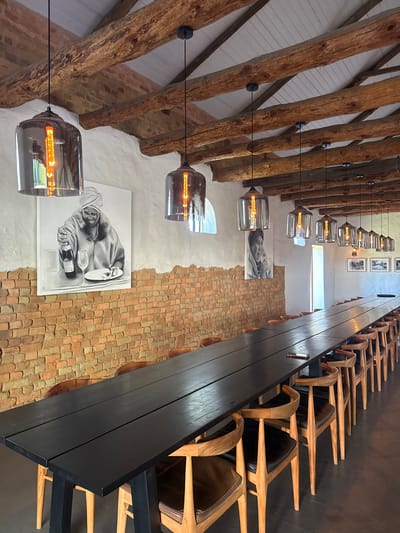 A high-angle interior shot shows a restaurant setting, featuring a long, black wooden table lined with wooden chairs. The floor is brick. The wall has a white portion with black-and-white photographs or drawings displayed. Exposed wooden beams cross the ceiling, and hanging from the beams are pendant lights with illuminated filaments. Pictures can be seen on the wall above the long table. The location is in the Stellenbosch Local Municipality, South Africa.