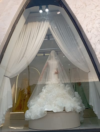 The image shows a wedding dress displayed in a shop window. The dress is white and features a voluminous skirt adorned with ruffled flowers. A sheer veil covers the mannequin wearing the dress, partially obscuring its features. The display is enhanced by flowing white curtains that frame the dress, creating a sense of elegance and drama. Behind the dress, a mannequin arm is visible, positioned as if adjusting the veil. Further in the background, a green fabric and a golden-colored material are visible, adding depth and color to the scene. Two spotlights illuminate the display from above. The shop window reflects the interior of the store and possibly the street outside, though these reflections are not clear enough to discern specific details. The overall impression is a luxurious and romantic presentation of a bridal gown. The setting appears to be a retail store, likely a bridal boutique. The lighting suggests it could be daytime, possibly afternoon, given the brightness. There is no visible text in the image.