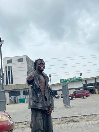A young Black man with braided hair and an open gray jacket stands in a paved outdoor area. He is smiling and pointing his left index finger towards the camera. The building behind him has the text "PRINCE EBANO SUPERMARKET" in green lettering on a white background. Several cars are visible in the parking lot of the supermarket, including a red minivan and a dark sedan. The sky is overcast. The scene appears to be in a commercial area, possibly in the city of Ojomu, Nigeria.