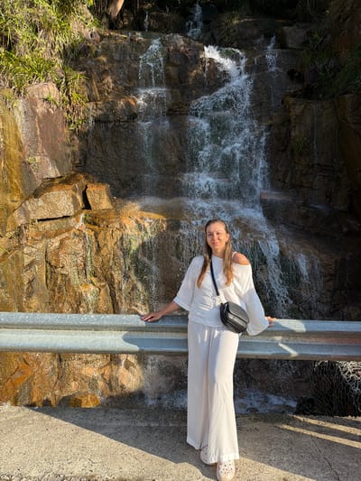 A woman stands in front of a waterfall, leaning on a metal guardrail. The waterfall cascades down a rocky cliff face with patches of green vegetation. The woman is wearing a white off-the-shoulder top and matching wide-leg pants. She has a black crossbody bag slung over her shoulder. Her feet are clad in light-colored Crocs. The ground in front of the guardrail is paved with concrete, and a long shadow stretches across it from the right side of the frame. The scene appears to be outdoors, likely in a natural setting within or near the city of Nam Khanh Vinh, Vietnam.