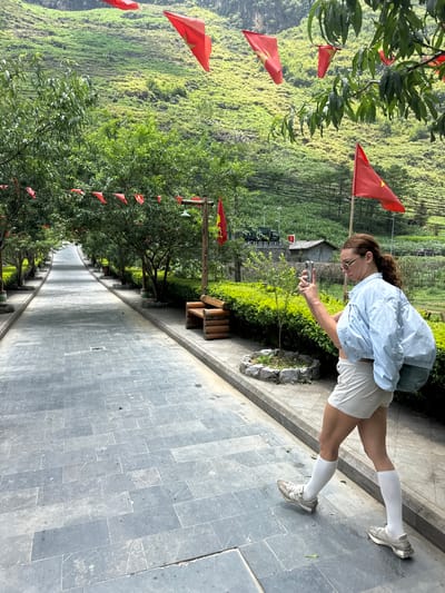 A woman in light blue jacket, white shorts and white knee-high socks stands on a stone-paved pathway, holding a phone. The pathway is lined with trees adorned with small red flags. A steep, green hillside rises to the left of the pathway. The scene is located in Mèo Vạc Commune, Vietnam.