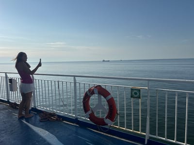 A woman stands on the deck of a ferry, holding a phone up to take a picture. She is wearing a pink patterned tank top and white shorts, and her hair is blowing in the wind. The deck of the ferry is blue with some white railings. A life preserver is hanging on the railing, with "ROBIN HOOD" and "EMDEN" visible on it. In the background, the sea stretches out to the horizon under a clear blue sky with a few wispy clouds. Several ships are visible in the distance. The sun appears to be low in the sky, suggesting it is either early morning or late afternoon. The overall mood is peaceful and scenic.