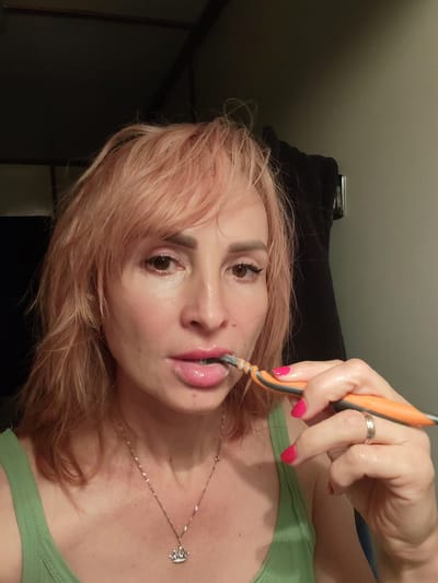 A woman with pink-tinted hair is brushing her teeth with an orange and gray toothbrush. She is wearing a green tank top and a silver necklace with a crown pendant. She has bright pink nail polish on her fingernails and a silver ring on her left hand. The background appears to be a bathroom, with a dark wall and a towel hanging on a hook. The lighting is dim. There is no information in the image that indicates the location in the city of Durasovo, Russia.
