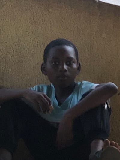 A child, presumably a boy, is seated. He appears to be eating something held in his right hand. He has a short haircut, dark skin, and is wearing a light blue shirt and dark shorts. He is facing the camera. The background is a textured, yellow surface. The photograph is taken at an angle, with the subject slightly tilted. The lighting is somewhat dim, casting shadows across the image.