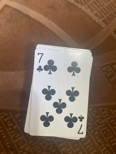 The image shows a deck of playing cards, specifically the Seven of Clubs, fanned out on a patterned brown surface. The cards are white with black pips and numbers. The top card clearly displays a "7" in the top left and bottom right corners, with multiple club symbols scattered across its face. The other cards in the deck are partially visible, suggesting they are also Seven of Clubs cards, as indicated by the visible club symbols. The background surface has a textured pattern. The lighting suggests it is indoors, with no indication of time of day or weather. The image does not depict people or any specific activity or event beyond the arrangement of the cards. The location context provided is Esik, Kazakhstan.