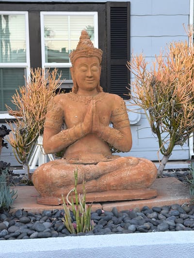 A terra-cotta statue of a meditating Buddha sits outside a building with light blue siding and dark brown trim. The Buddha is depicted in a seated position with hands pressed together in front of its chest, wearing an ornate crown. The statue is placed on a concrete base, surrounded by a bed of dark grey river rocks. Two sparsely branched, coral-like plants, one on each side of the Buddha, add a touch of color with their reddish-orange stems. In the background, there are two white-framed windows with horizontal blinds. The overall scene suggests an outdoor decorative element in a residential or commercial setting.