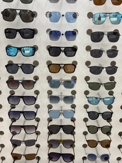 This is a close-up, high-angle shot of a display of sunglasses in a store. The sunglasses are arranged in rows on a white pegboard. There are many pairs of sunglasses, with frames of various colors and materials, and lenses of different tints and shades. Some of the lenses are reflective, showing distorted images of the store interior. The overall impression is one of abundance and variety, typical of a retail environment. The lighting is bright and even, suggesting an indoor setting. The image is static, with no people or action visible, focusing solely on the product display. The scene is likely a sunglasses shop, possibly in a tourist destination like Saint-Tropez, France, given the context provided. The time of day and weather are not discernible from the image. There is no visible text in the image.