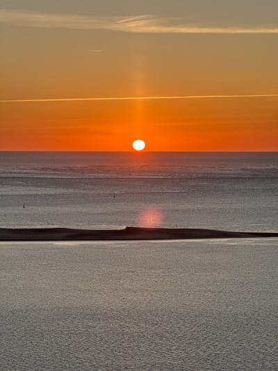 This is a vertical, eye-level, full shot of a sunset over the ocean, likely the Atlantic Ocean off the coast of La Teste-de-Buch, France. The sun, a bright orange disc, is positioned just above the horizon, casting a vibrant orange glow across the sky and a shimmering reflection on the water. The sky transitions from a pale yellow near the horizon to a dusty blue at the top, with thin, wispy clouds and two distinct contrails from aircraft visible.
The ocean is divided into three distinct horizontal bands. The uppermost band, closest to the horizon, is a calm, dark gray expanse with subtle ripples. Two small, dark buoys are visible in this section. Below this is a slightly lighter gray band, also calm, with a faint reflection of the sun's light. The lowest band, occupying the bottom half of the image, is a muted gray, textured with small, choppy waves that catch the light.
In the foreground, a dark, low-lying landmass, possibly sand dunes or a very low island, separates the upper two water bands from the bottom one. Its surface is mostly in shadow, but a thin line of bright light runs along its top edge, reflecting the setting sun. The overall atmosphere is serene and tranquil, typical of a sunset scene over a coastal environment. There are no people or any other discernible subjects or actions within the frame.