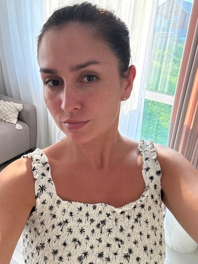 The image shows a woman with dark hair pulled back, wearing a white top with a black palm tree print. She has freckles on her face and is wearing a small diamond stud earring in her left ear. The background features sheer white curtains and a window with a view of a building and greenery. A grey sofa with a textured throw pillow is visible to the left. The lighting suggests it is daytime, possibly indoors with natural light filtering through the window. The location is noted as Nizhnyarkeyevo, Russia.